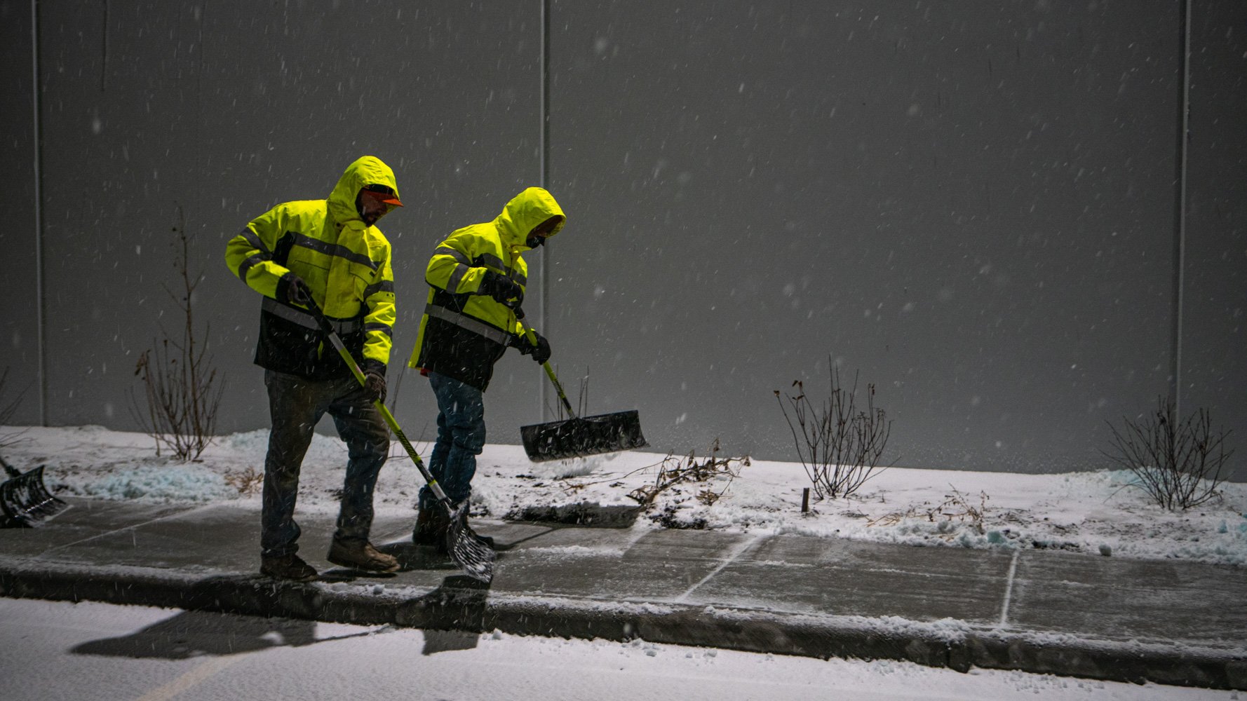 commercial snow removal team shoveling the sidewalk at an industrial facility at night