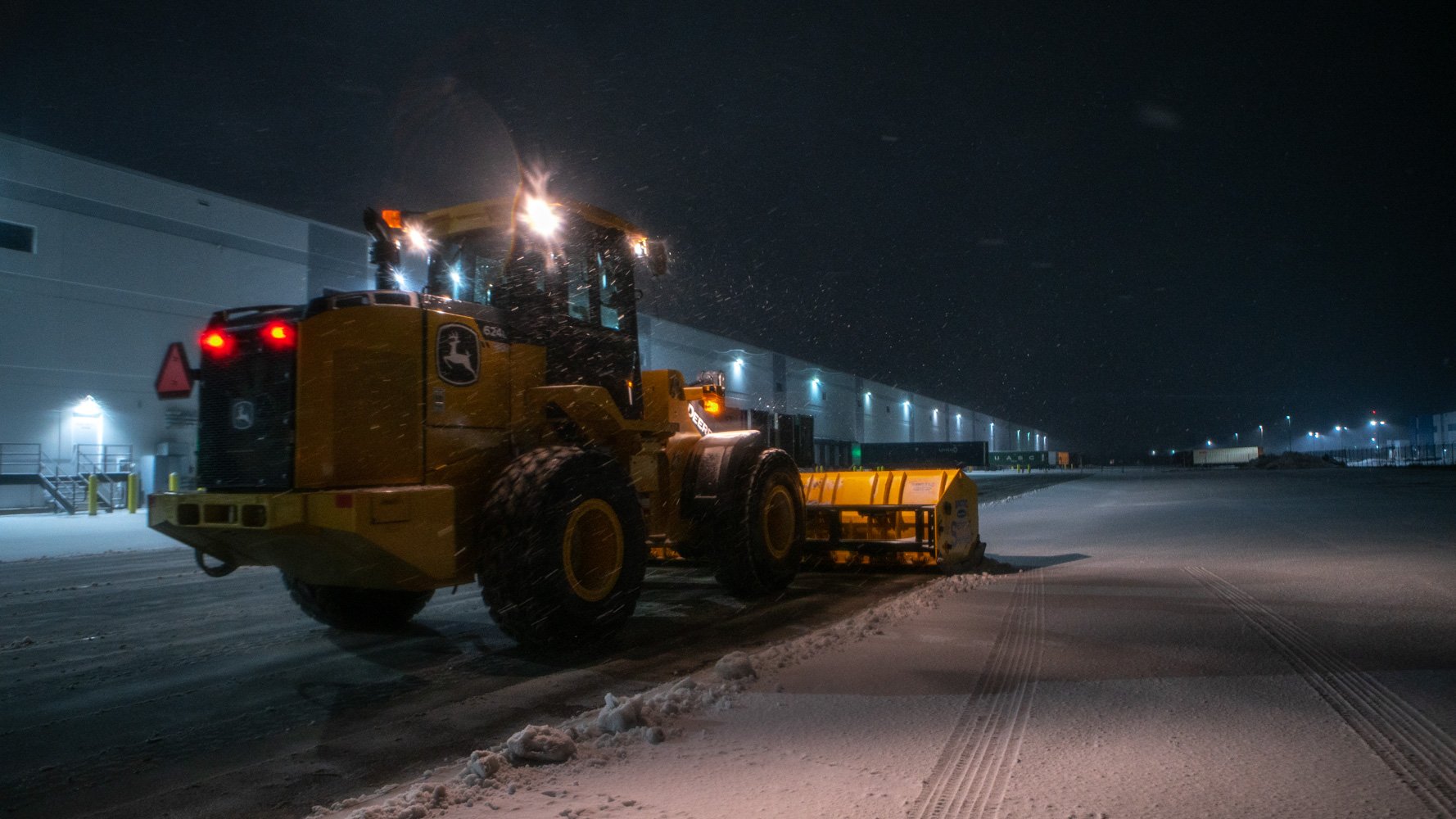 commercial snow loader plowing a warehouse parking lot at night